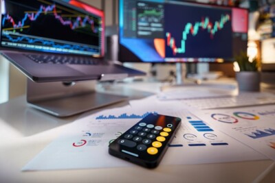 o que é um home broker: Calculadora de smartphone na mesa com gráficos financeiros atrás